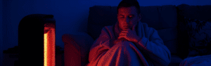 Distressed man sitting on a couch, bundled up next to a heater.