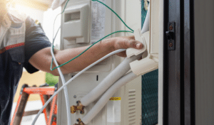 Technician installing an outdoor air conditioning unit with insulated pipes and electrical wiring.