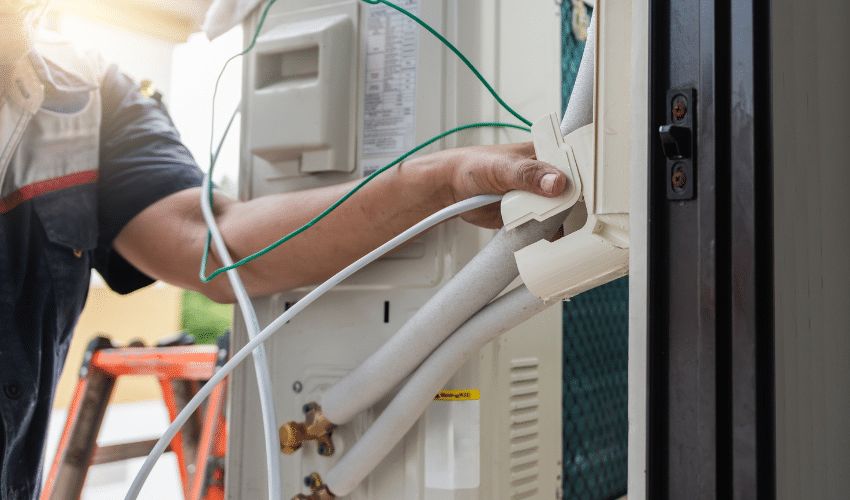 Technician installing an outdoor air conditioning unit with insulated pipes and electrical wiring.