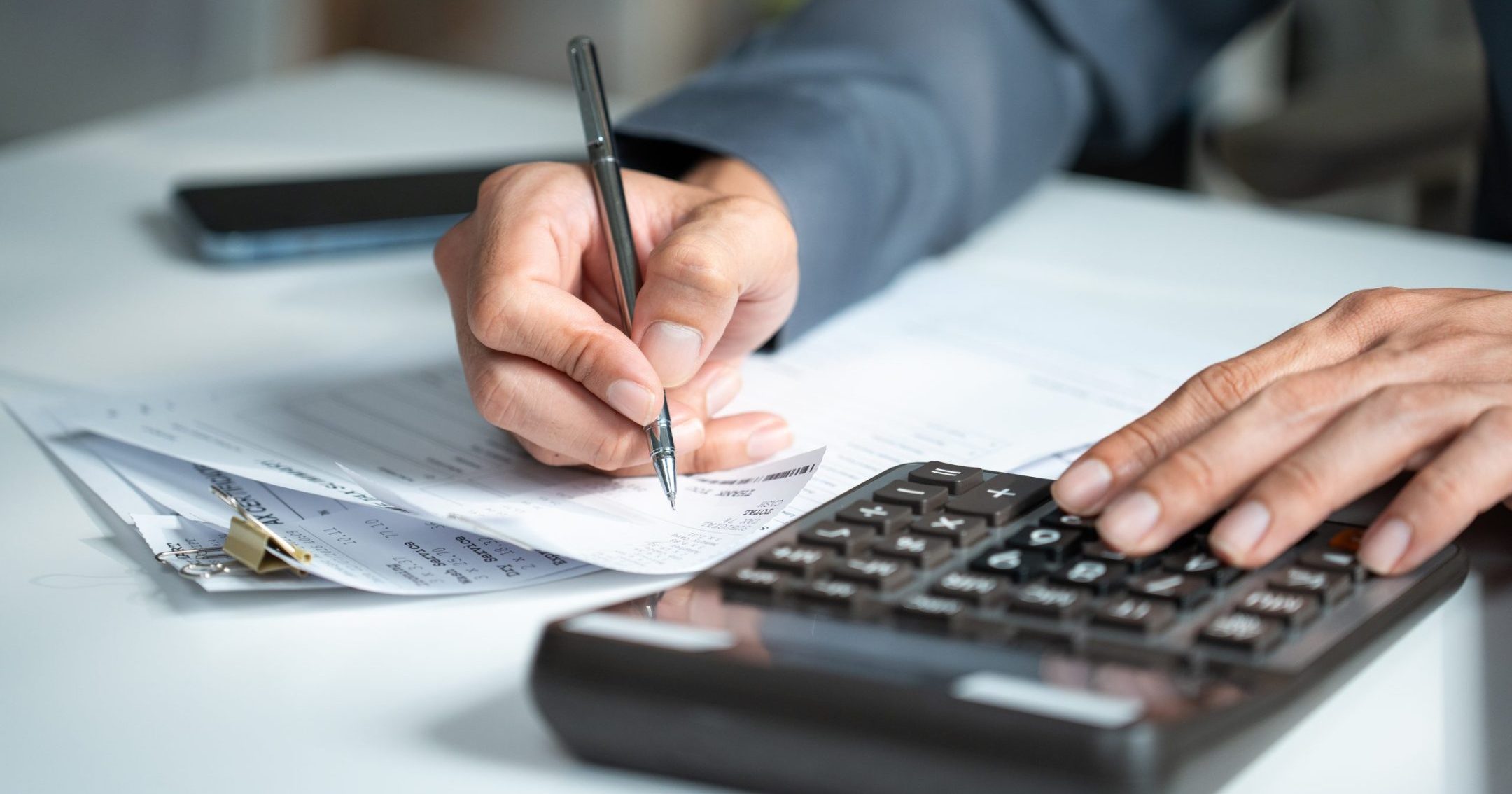 Accountant hand using a calculator and writing on financial expense receipt documents at an office desk, representing tax calculation and auditing